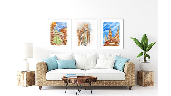 Set of three atmospheric American Southwest watercolor canvas prints from the American Escape collection. Psychedelic Sedona Red Rocks Southwestern wall art watercolor Canvas Print. Stylized American Southwest watercolor canvas print. Watercolor paintings archival prints against a white wall, hanging above a rattan sofa in a living room decor.  Features Adobe Abode canvas print and Land Enchanted canvas print from the American Escape collection. 